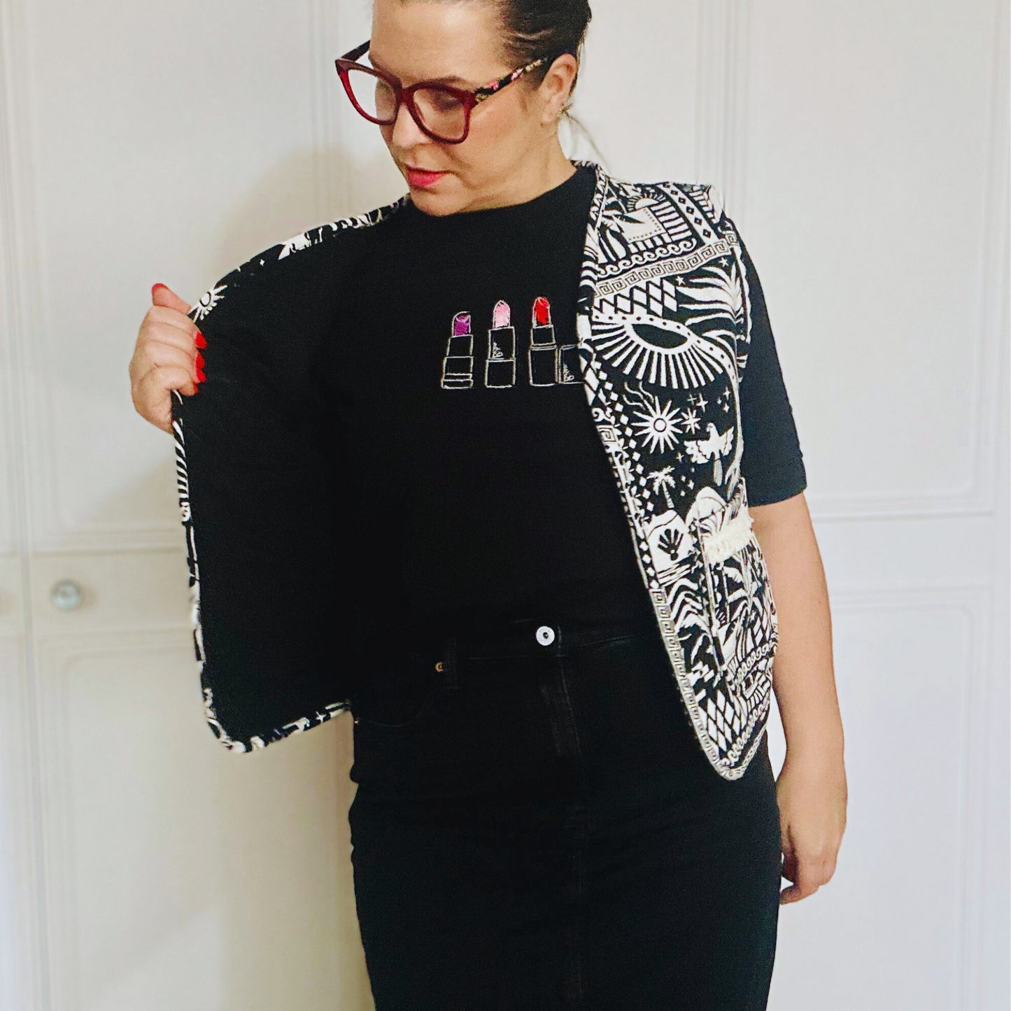 Awoman with dark hair faces the camera wearing a Black and white patterned vest. The vest features intricate designs, including geometric shapes, birds, and plants, with two front pockets that match the detailed print. The bold design gives the vest a unique, handcrafted look.