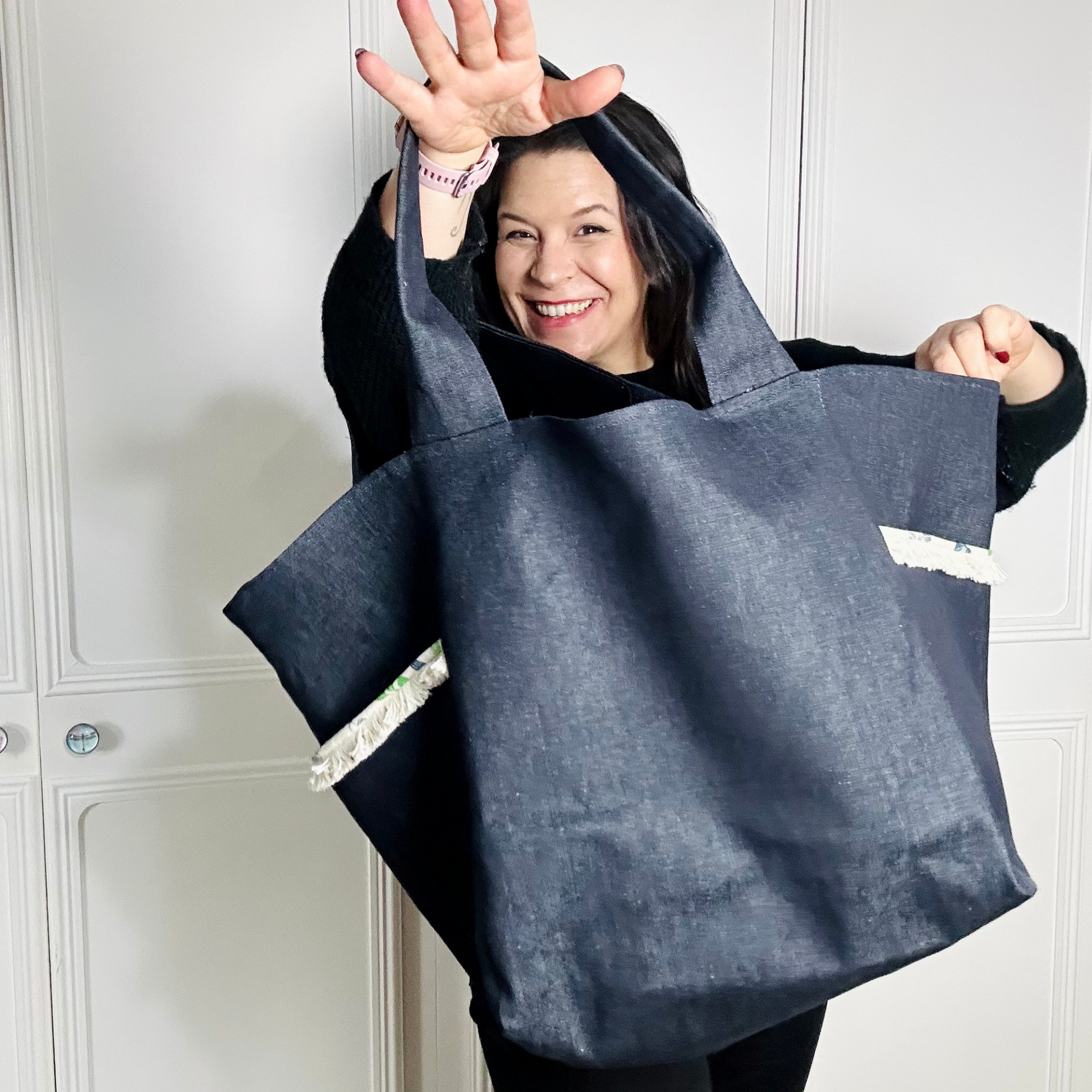 A woman wearing all black is facing the camera against a white background. She is holding up a large denim bag and smiling through the bag handle hole.