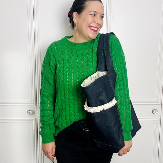 A woman stands again a white background wearing a green jumper looking over her shoulder to the left. She has brown hair and is carrying a large denim bag