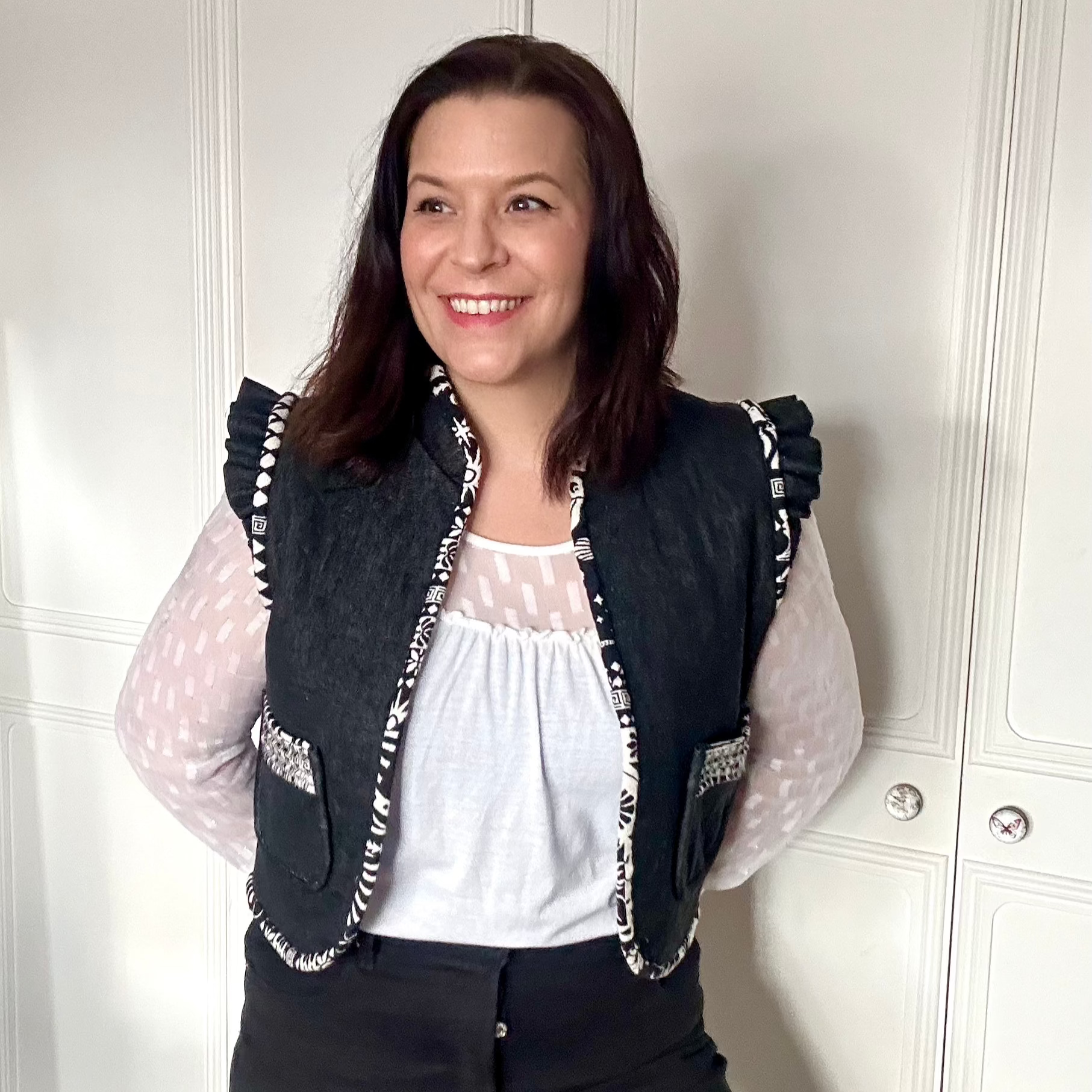 A white woman faces the camera wearing a black waistcoat with black and white trim. Her hands are behind her back and she stands in front of a white wall.