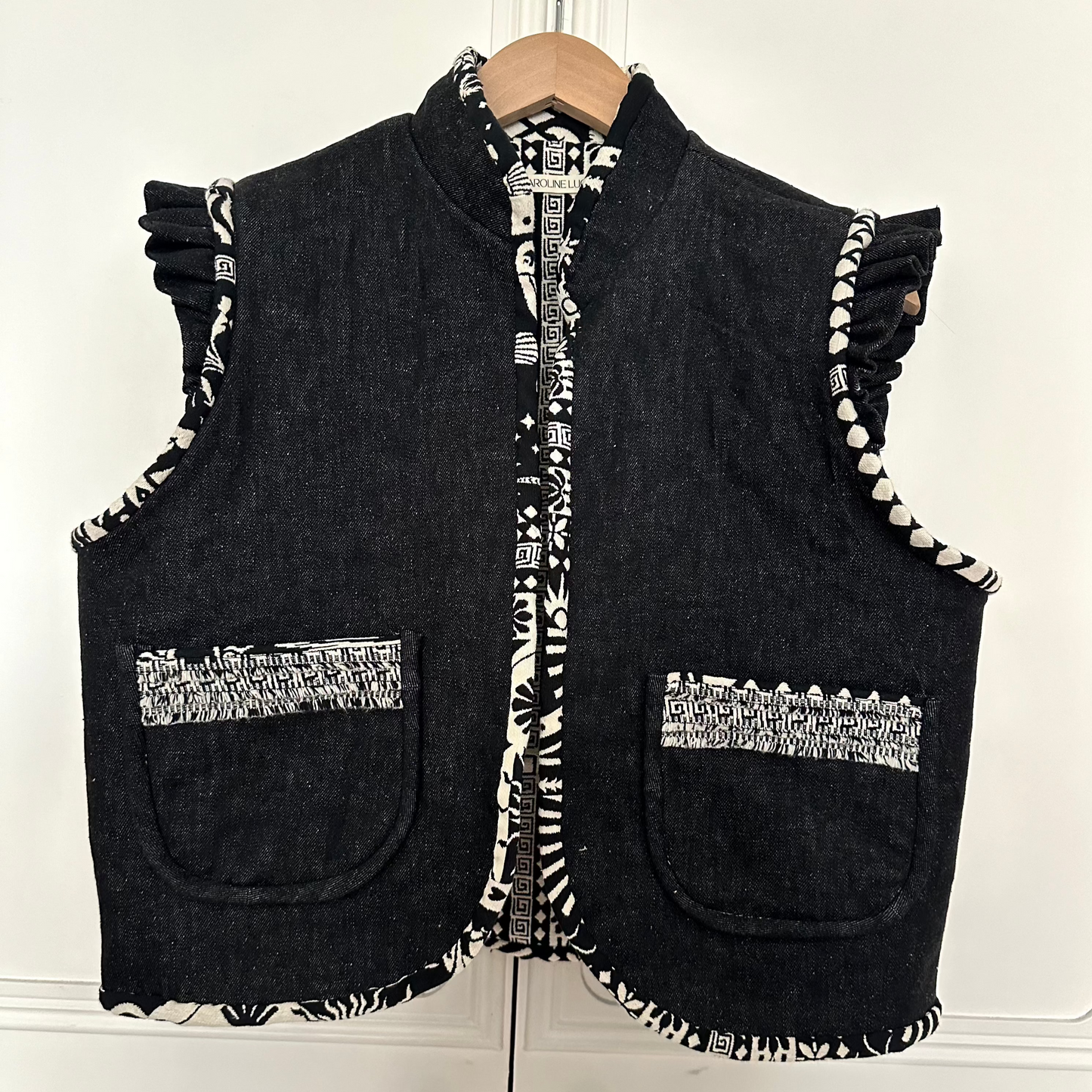 A black denim waistcoat hangs on a hanger against a white background