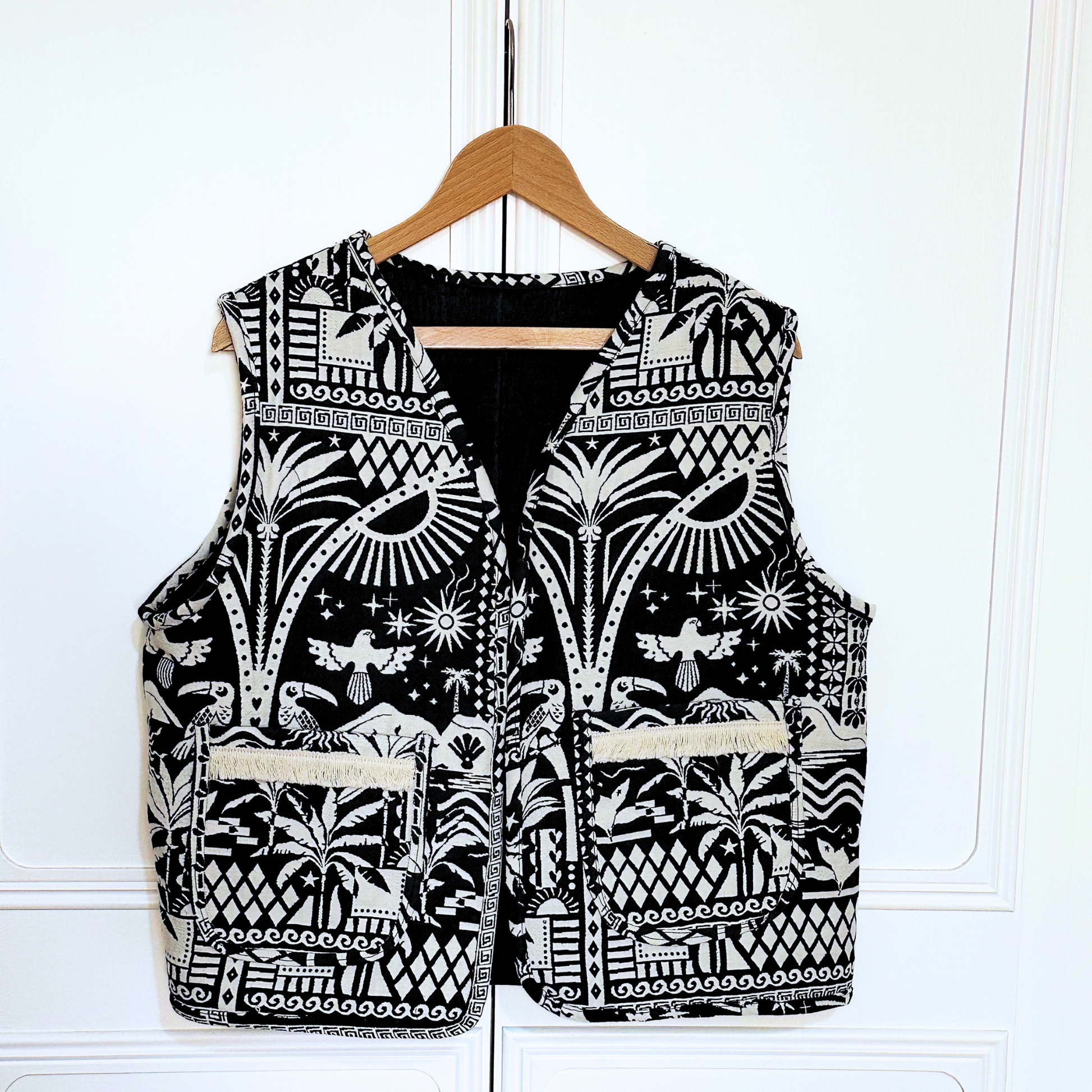 Black and white patterned vest hanging on a wooden hanger against a light-coloured background. The vest features intricate designs, including geometric shapes, birds, and plants, with two front pockets that match the detailed print. The bold design gives the vest a unique, handcrafted look.