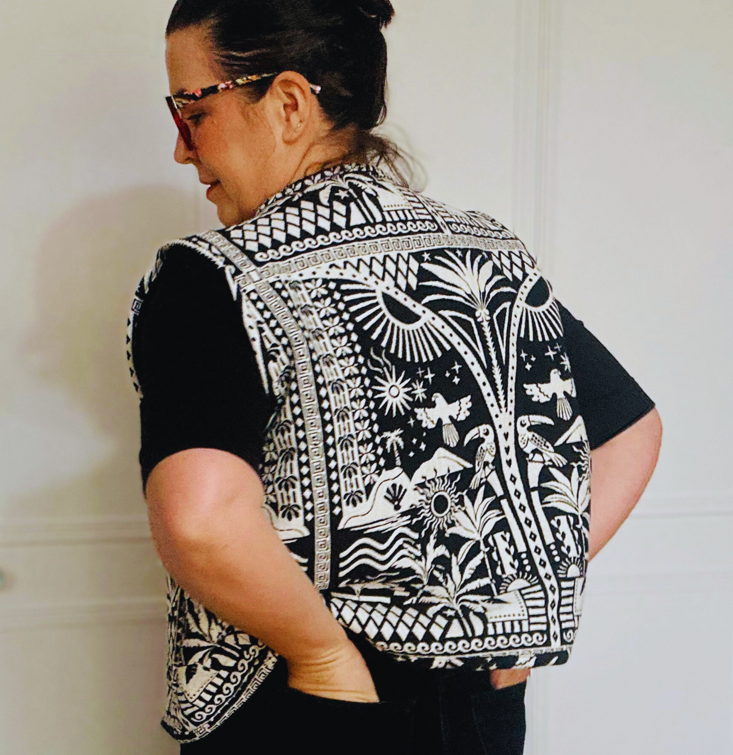 woman with dark hair  with her back to csamera  wearing a Black and white patterned vest. The vest features intricate designs, including geometric shapes, birds, and plants, with two front pockets that match the detailed print. The bold design gives the vest a unique, handcrafted look.