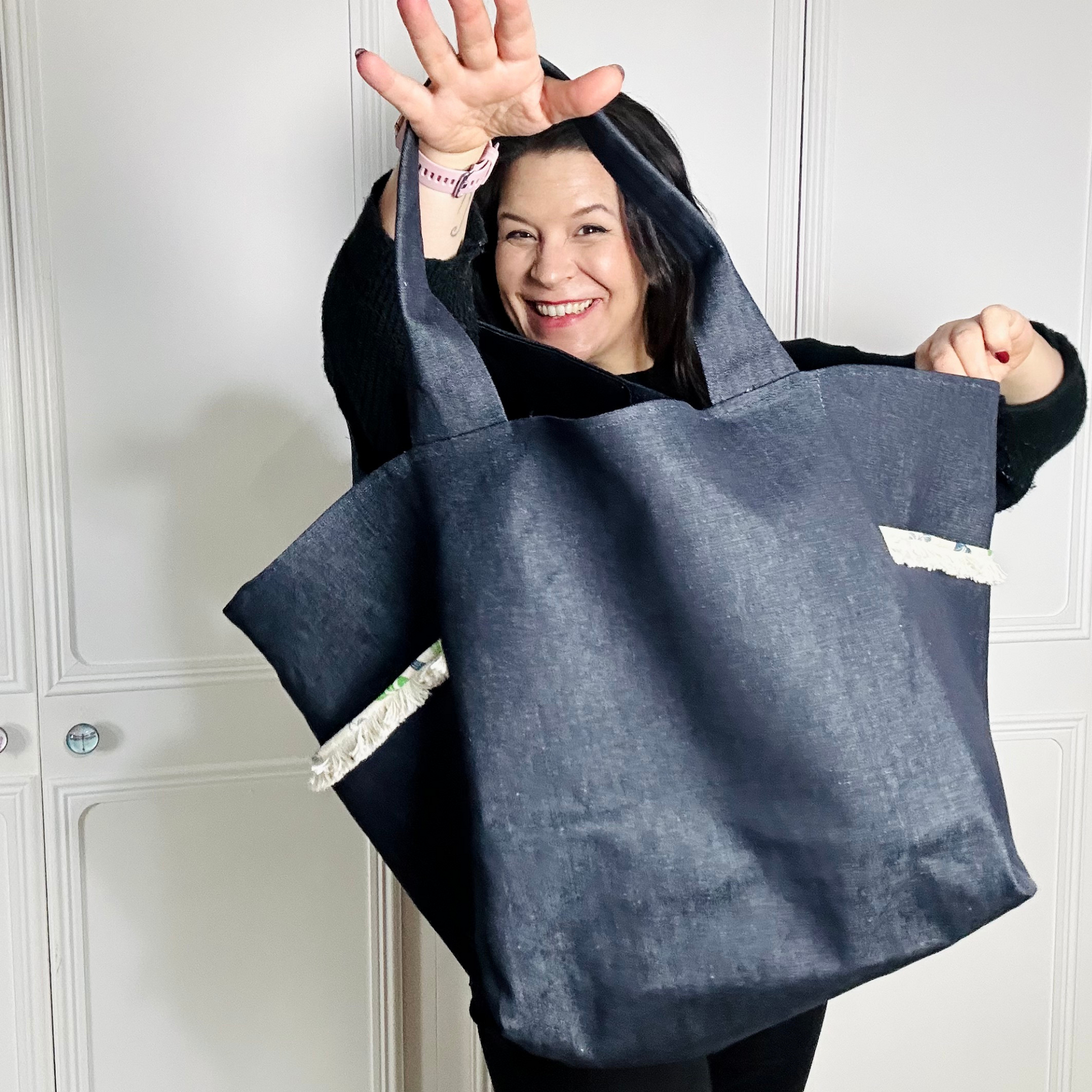 A woman wearing all black is facing the camera against a white background. She is holding up a large denim bag and smiling through the bag handle hole. 